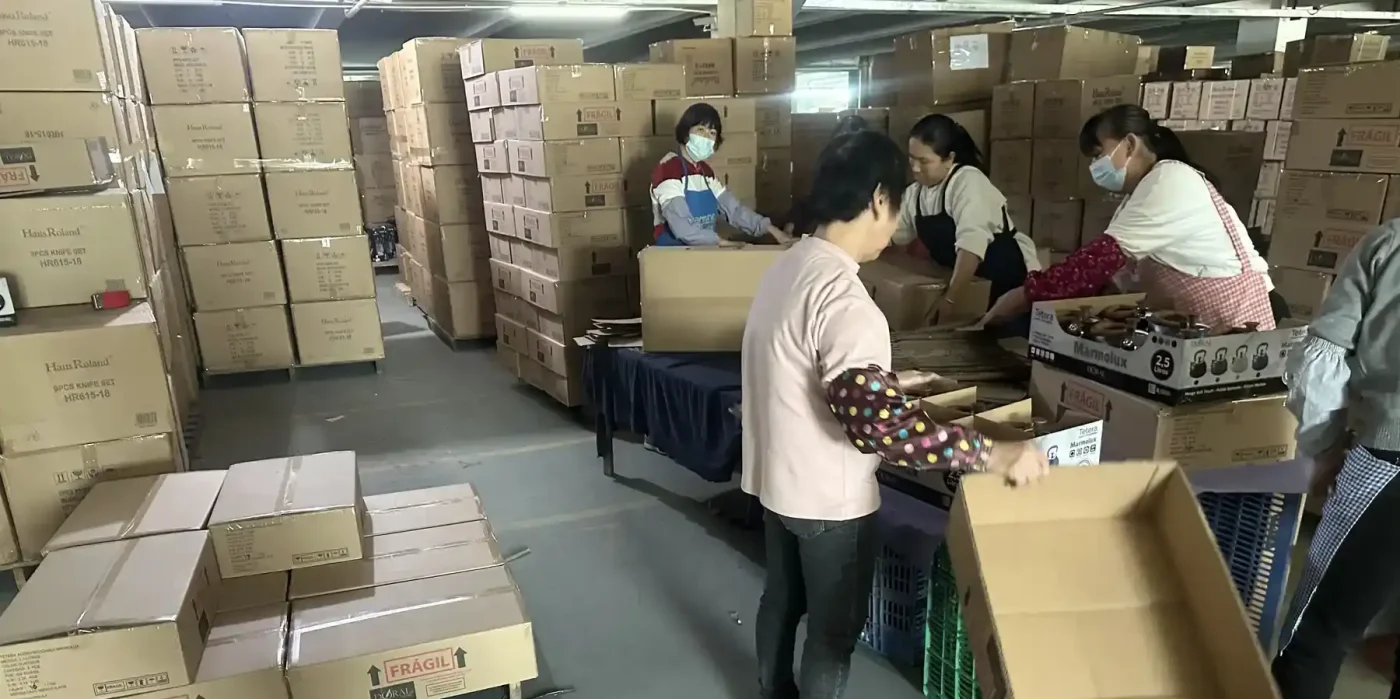Workers preparing cartons in a cookware packing area with stacked labeled boxes